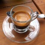 A single shot of expresso in a clear glass with a saucer and spoon