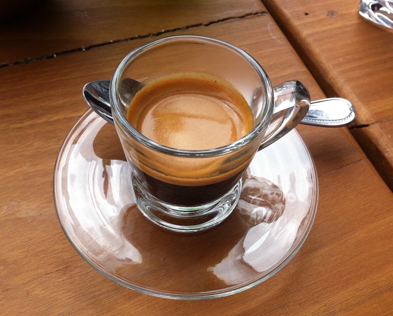 A single shot of expresso in a clear glass with a saucer and spoon